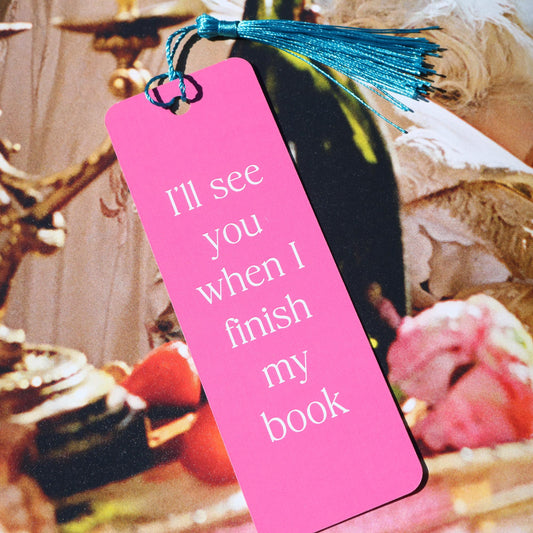A pink paper bookmark with a tassel, featuring the text 'I'll See You When I Finish My Book' from a book.