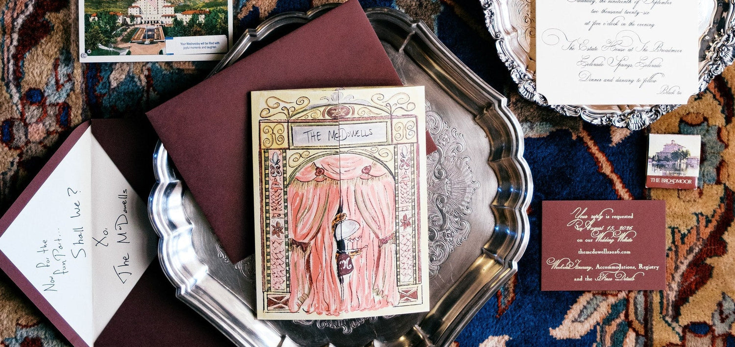 An elegant vintage-style wedding invitation suite arranged on a richly patterned blue and brown oriental rug. The suite includes an illustrated wedding program, an invitation card with a pink curtain, a silver charger plate, a small postcard, a white envelope with handwritten text, and other coordinating stationery pieces.