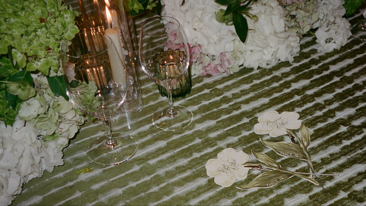A close-up of a table setting featuring a green and white striped tablecloth, clear glass candle holders with lit taper candles, white and pale pink hydrangea flowers, and a single white flower with gold leaves lying on the table.